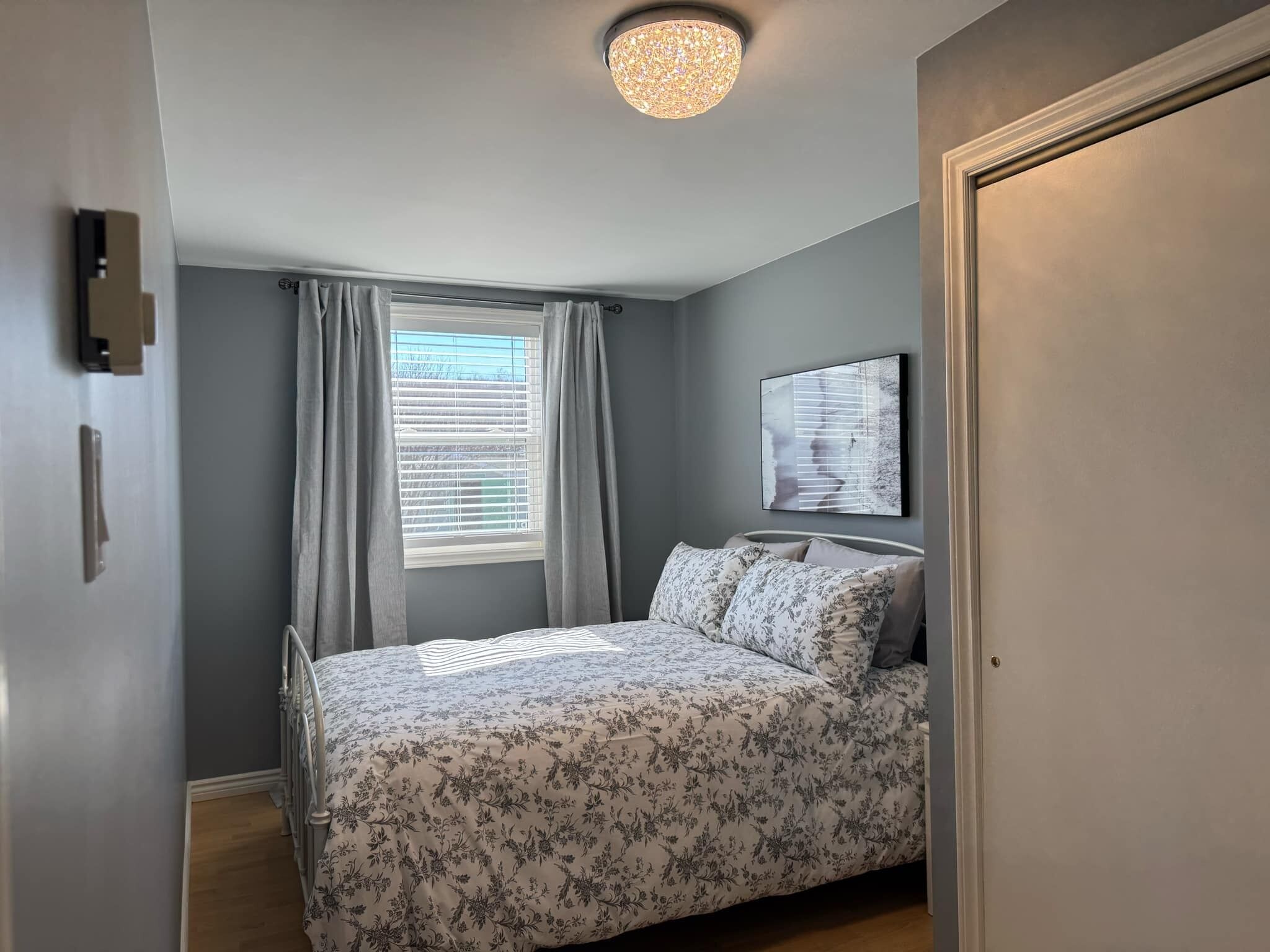 A bedroom with a bed, a window with curtains, a ceiling light, and a wall-mounted artwork.