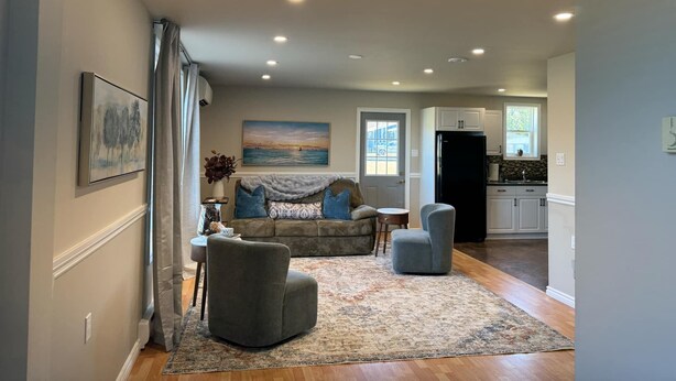 A spacious living area with a sofa, two armchairs, a coffee table, and a rug. There is a painting on the wall, a window with curtains, and a kitchen in the background.