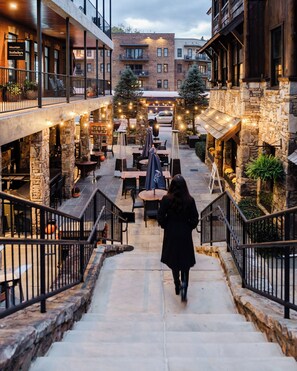 A person descending a stone staircase in a building with multiple floors, leading to an outdoor seating area with tables and chairs, surrounded by stone walls and string lights. - Blue’s Nook: Mountain View’s, Cedar Barrel Hot Tub, and close to Downtown (Blue Ridge)