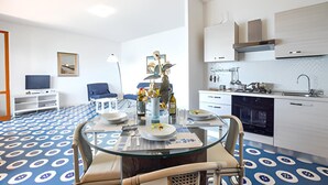 A modern room with a glass dining table, blue patterned carpet, white walls, a TV, a chair, and a kitchen area with white cabinets and a sink.
