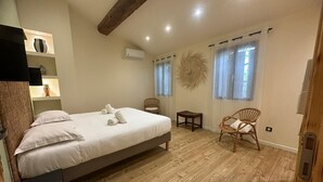 A bedroom with a wooden floor, a bed with white linens, two wicker chairs, a small table, and a wall-mounted air conditioner. - La villa verte Domaine de la tapie de Bouchet - in the Camargue (Arles)
