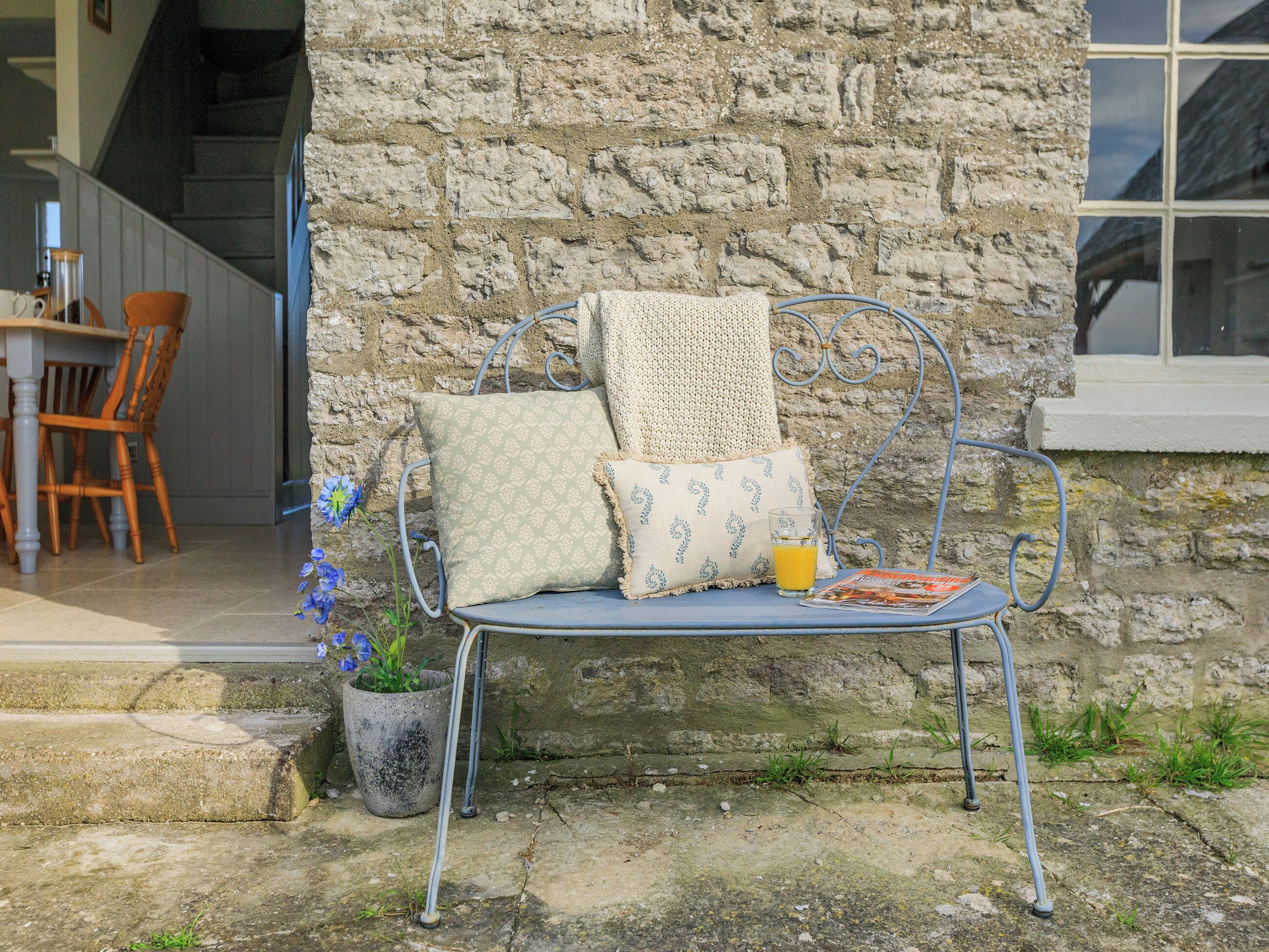 A metal bench with cushions against a stone wall, a potted plant, and a glass of orange juice on the ground.