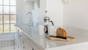 A modern kitchen with a marble countertop, a coffee maker, a loaf of bread, and a window with a view of trees. - The Cottage at Brook Farm (Southwood, near Glastonbury)
