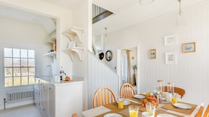 A bright kitchen with white cabinetry, a dining area with wooden chairs and a table set for breakfast, and a staircase leading to another level. - The Cottage at Brook Farm (Southwood, near Glastonbury)