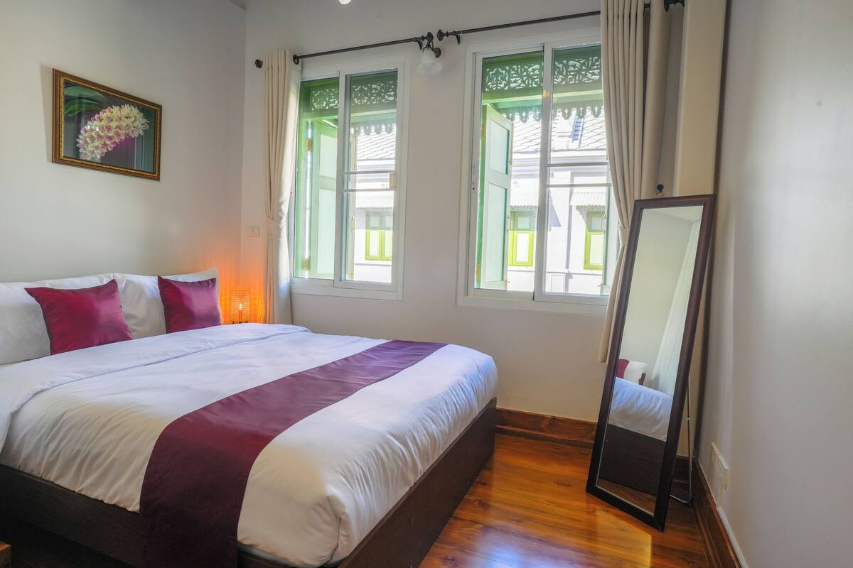 A neatly made bed with a white and maroon bedspread, a large mirror, and a window with green shutters.