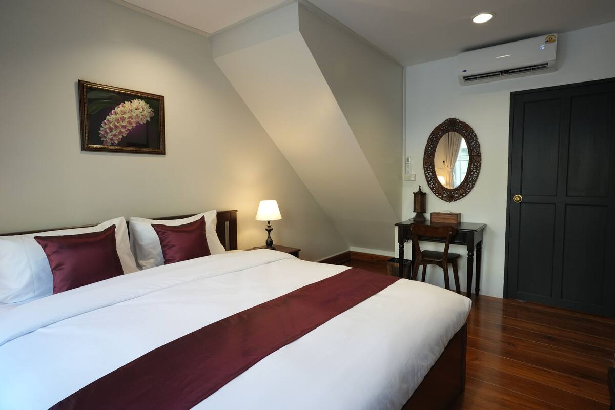 A bedroom with a large bed, a bedside table with a lamp, a wooden desk, a mirror, and an air conditioning unit.