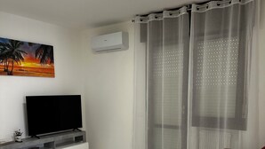Un salón con televisor de pantalla plana, una mesa de centro blanca, un sofá beige y cortinas ligeras.