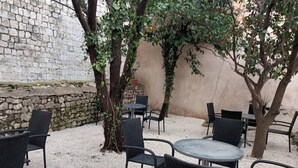 An outdoor seating area with wicker chairs and tables, surrounded by trees and a stone wall. - Three-bedroom apartment with balcony (Dubrovnik)