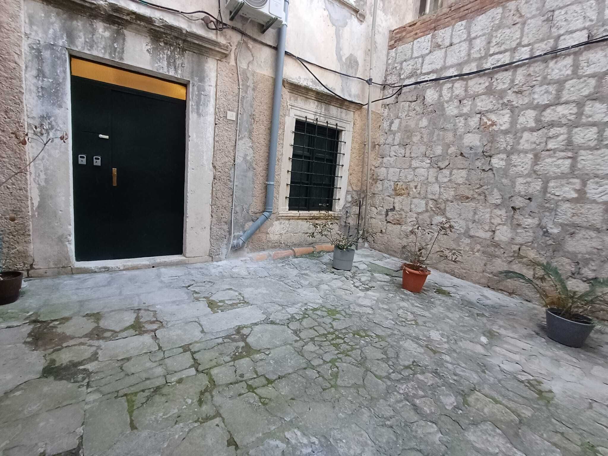 A courtyard with a stone floor, a black door, a window with metal bars, and potted plants.