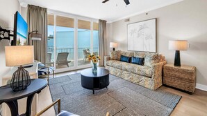 Un salon moderne avec un canapé, une table basse et des lampes. On y trouve une grande fenêtre avec des rideaux, un ventilateur de plafond et une œuvre d’art encadrée au mur.