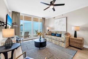 Un salon moderne avec un canapé, une table basse et des lampes. On y trouve une grande fenêtre avec des rideaux, un ventilateur de plafond et une œuvre d’art encadrée au mur.
