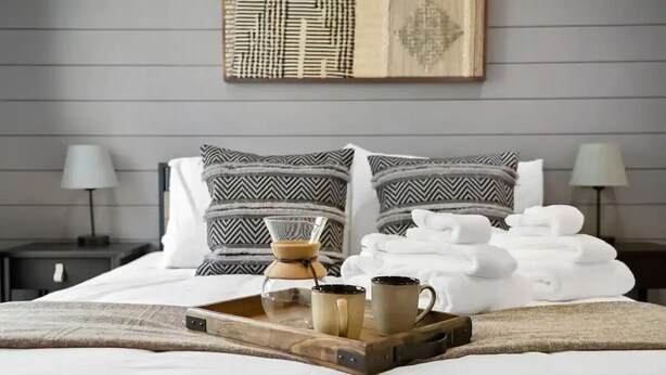Un lit bien fait, agrémenté d’oreillers à motifs, un plateau avec des tasses et une cafetière, et des serviettes pliées.