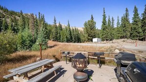 Un terrain de camping équipé de tables de pique-nique, d’un foyer et d’un espace barbecue, le tout entouré d’arbres et adossé à une colline.