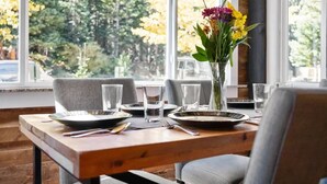Une table à manger dressée avec des assiettes, des couverts et des verres, et un vase de fleurs posé sur le rebord de la fenêtre.