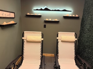 Ein Wellnessraum mit zwei Liegestühlen, einem wandmontierten Regal mit Kristallen, einer Wandleuchte und einer grünen Wand.