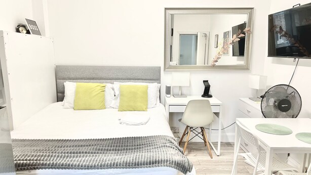 A hotel room with a bed, a desk, a chair, a mirror, a TV, and a fan. - Flat 2  (London)