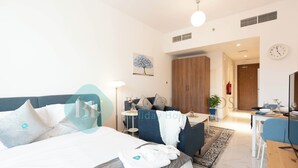 A modern hotel room with a large bed, a desk, a dining area, and a view of the hallway. - Bloomfields Elegant STU in Oasis Masdar (Abu Dhabi)