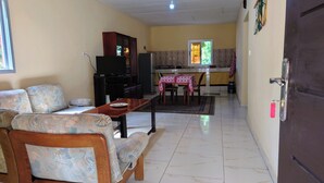 A living room with a sofa, a coffee table, and a TV stand. There is a kitchen area with a dining table and chairs. - Red Villa on Lionsrest - a tropical garden 500m from the Atlantic beach (Grand Bassam)