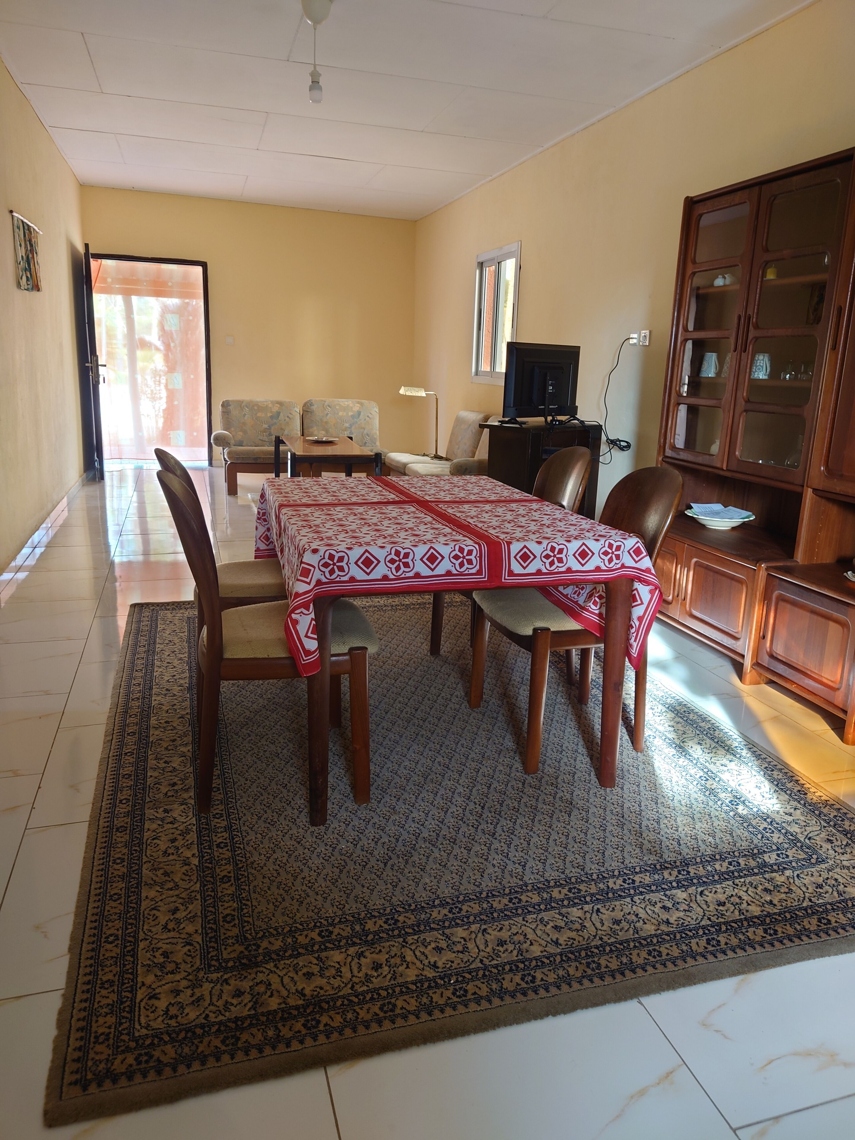 Un comedor con una mesa dispuesta para seis personas, rodeada de sillas. Un televisor está montado en la pared, y se ve un aparador de madera a la derecha.