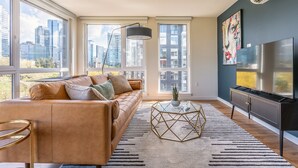 A modern living room with a leather sofa, a glass coffee table, a flat-screen TV, and large windows offering city views.