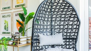 A black wicker chair with a white cushion, a gold cart with a plant, and framed cocktail posters on the wall.