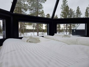Une chambre avec deux lits, du linge de lit blanc et une vue sur les arbres et un chalet, visible à travers de grandes fenêtres.