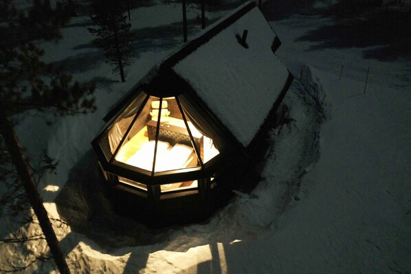 Une structure hexagonale recouverte de neige, dont l’intérieur est éclairé, et qui est entourée de neige et d’arbres.