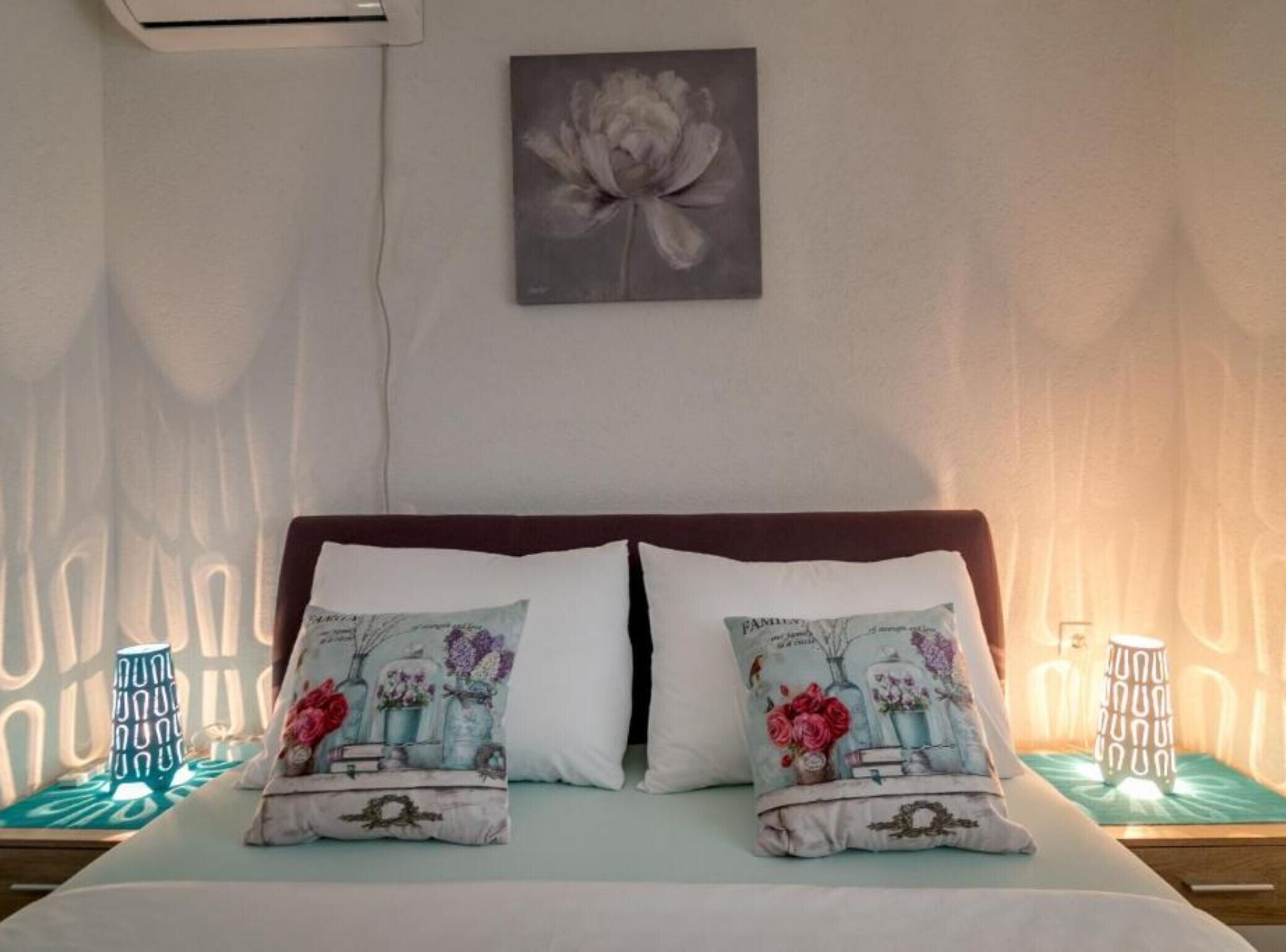 A bed with two decorative pillows, a wall-mounted air conditioner, and a floral painting above the bed.