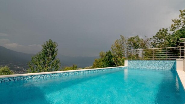 Ein klares, blaues Schwimmbecken mit einer gefliesten Kante, umgeben von Bäumen und Blick auf ferne Berge.