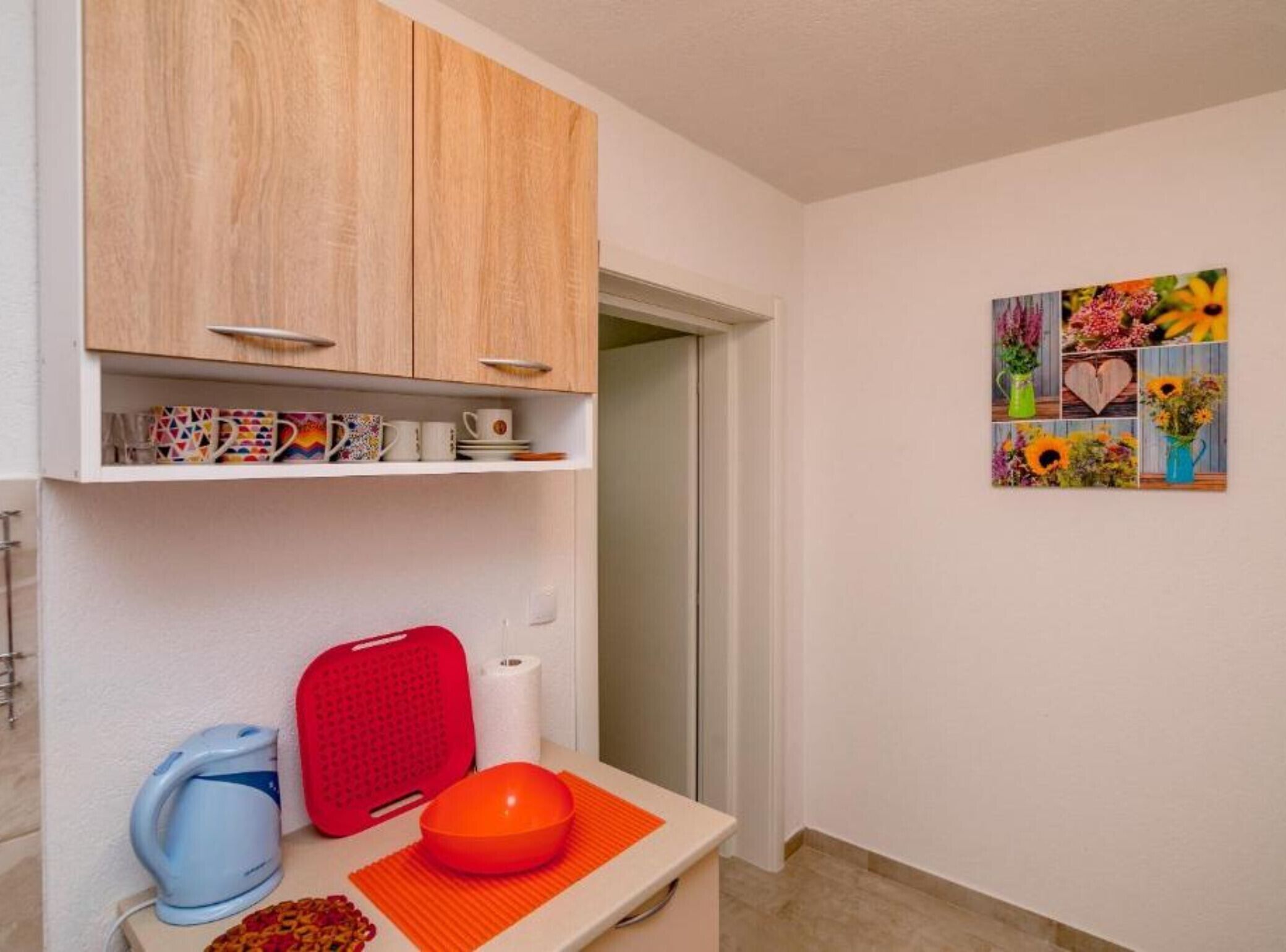 A small kitchen area with a wooden cabinet, a kettle, a red cutting board, and a bowl on a mat.