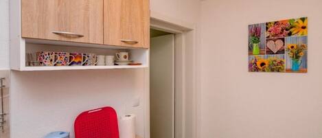 A small kitchen area with a wooden cabinet, a kettle, a red cutting board, and a bowl on a mat.