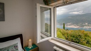 Ein Schlafzimmer mit einem großen Fenster und Blick auf eine Küstenlandschaft, einem Nachttisch mit Lampe und einem gerahmten Bild an der Wand.
