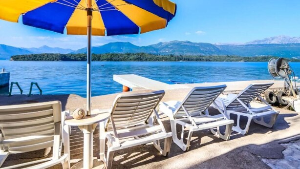 A lakeside setting with lounge chairs and a table under a striped umbrella, overlooking a calm lake and distant mountains. - Beachfront Escape – Steps from the Crystal-Clear Sea - One Bedroom Apartment (Bogisici)
