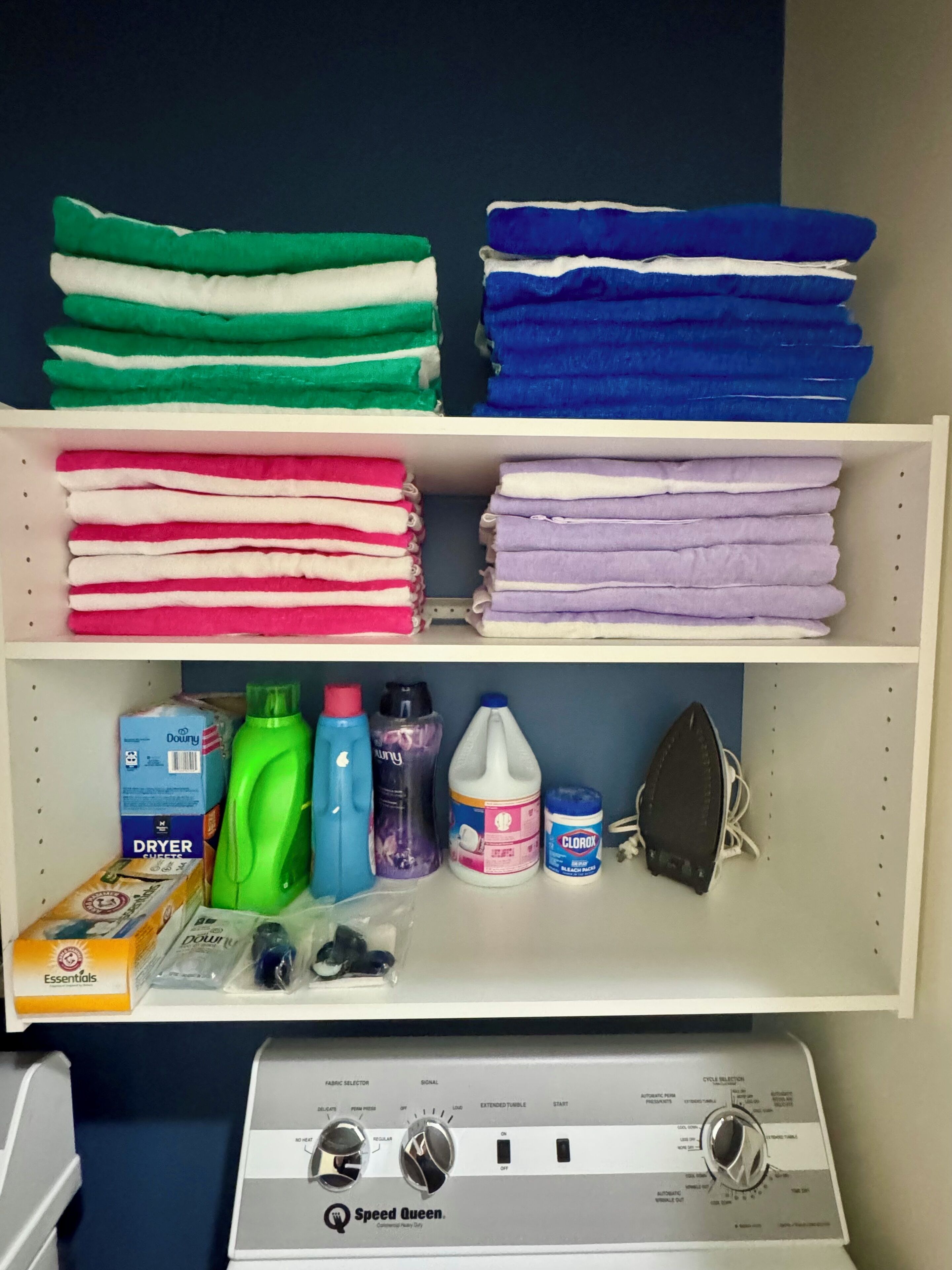 Un espace dédié à la buanderie, avec des étagères remplies de serviettes pliées de différentes couleurs et un sèche-linge doté d’un panneau de commande.