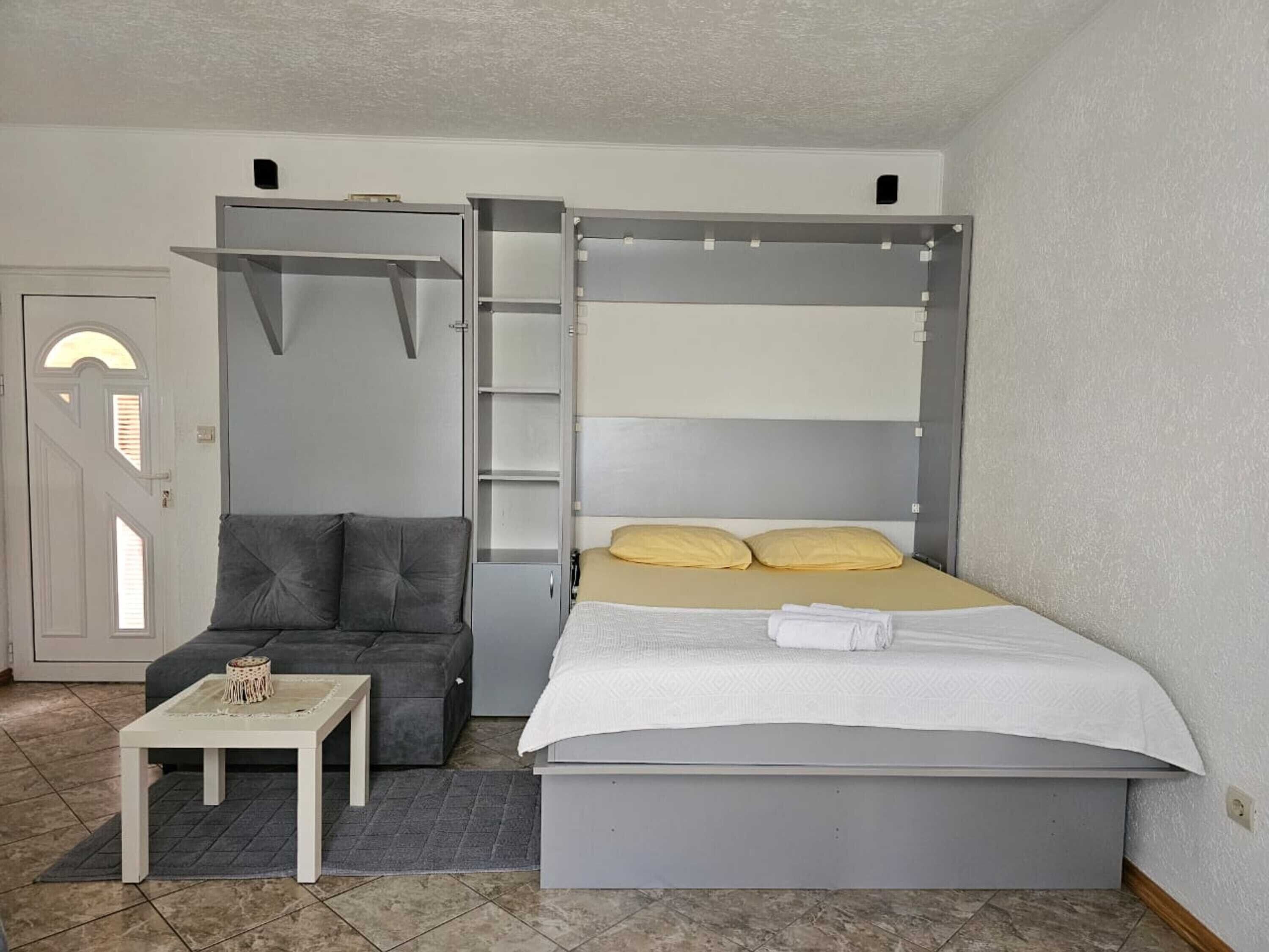 Ein kompakter Raum mit einem Bett, einem grauen Sofa, einem kleinen Tisch und einem Schrank mit Regalen.