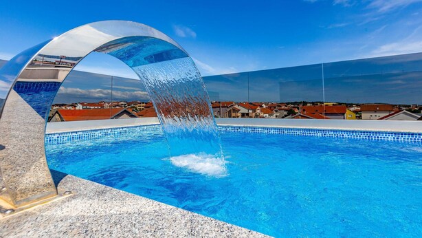A swimming pool with a clear blue water, a curved glass structure, and a view of houses in the background. - Three Bedroom Apartment with Balcony (Zadar)