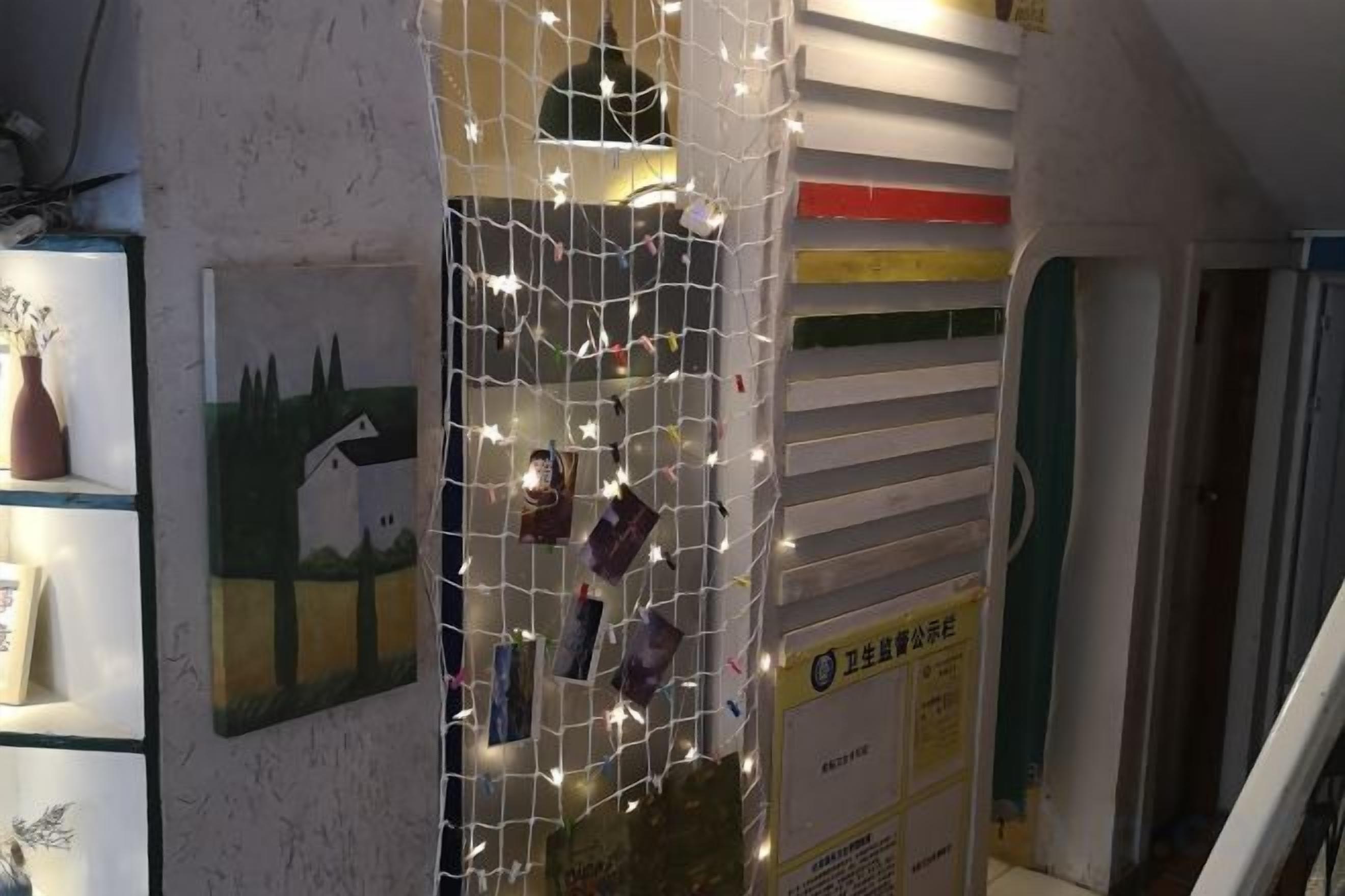 Een kamer met een muur versierd met lichtslingers en een schilderij. Er is een plank met een vaas en een fotolijst.