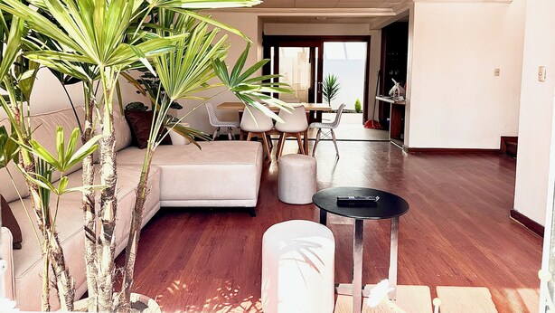 Un salón amplio con un sofá, una mesa de comedor y sillas. Varias plantas en macetas y una mesita con un control remoto.