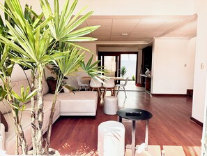 Un salón amplio con un sofá, una mesa de comedor y sillas. Varias plantas en macetas y una mesita con un control remoto.