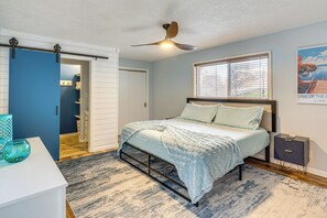 Ein Schlafzimmer mit einem Bett, einem Nachttisch, einem Deckenventilator, einem Fenster mit Jalousien und einer Tür zu einem Badezimmer.