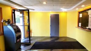 A hotel lobby with a yellow wall, a reception desk, an ATM, and a door.