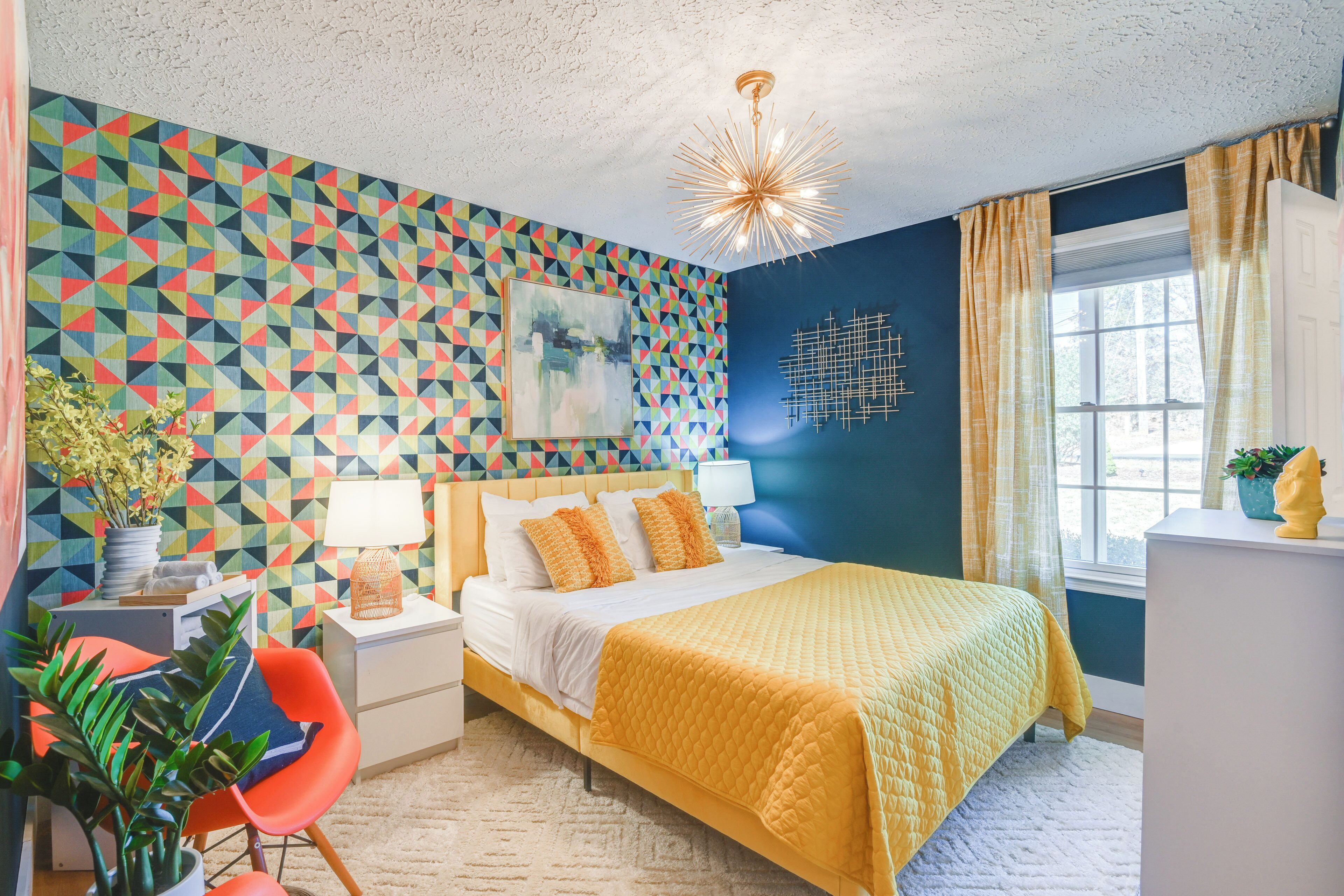 A bedroom with a geometric-patterned wall, a bed with a yellow quilt, a nightstand with a lamp, a chair, and a window with curtains.