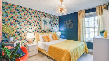 A bedroom with a geometric-patterned wall, a bed with a yellow quilt, a nightstand with a lamp, a chair, and a window with curtains.