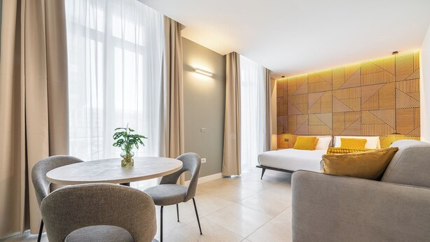 Ett modernt hotellrum med ett runt matbord, två stolar, en soffa och en säng med ett mönstrat sänggavel.