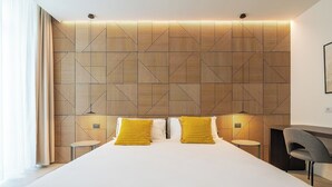 Ett modernt hotellrum med en sänggavel med geometriskt mönster, en stor säng med vita sängkläder, två sängbord med lampor och en grå fåtölj.