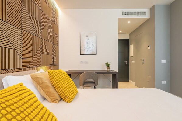A modern bedroom with a bed, a desk, and a chair. There is a wall with a geometric pattern and a framed picture on the wall.