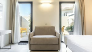 A hotel room with a sofa, a bed, a desk, and a view of the outside balcony. - JUNIOR SUITE DOUBLE BED (Cagliari)