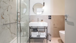 A modern bathroom with a glass shower enclosure, a white sink with a round mirror, and a toilet. - JUNIOR SUITE DOUBLE BED (Cagliari)
