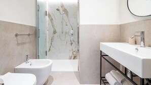 A modern bathroom with a glass-enclosed shower, a standalone bidet, and a vanity with a sink and towel rack. - Deluxe Double Room (Cagliari)