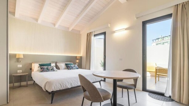 A modern hotel room with a large bed, a round dining table, two chairs, a small bedside table, and a sliding glass door leading to an outdoor area. - Deluxe Double Room (Cagliari)